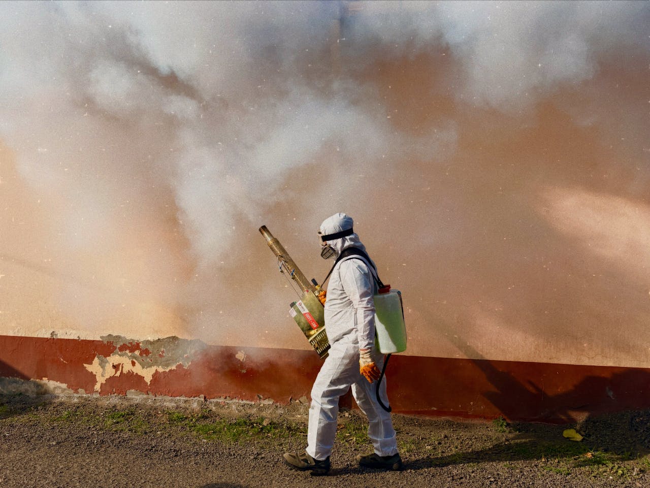 A worker in a full protective suit fumigates an area outdoors, spraying pesticide.
