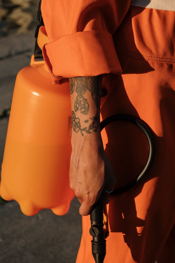 why-choose-us Person in orange coveralls holding a plastic sprayer, focus on tattooed arm.